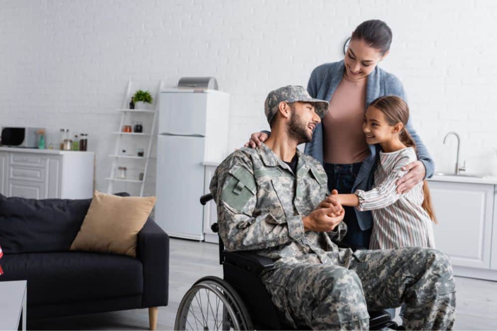 Smiling kid holding hand of dad in military uniform in wheelchair near mother at home